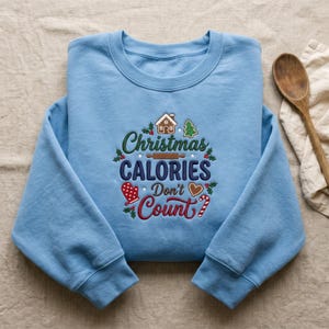 May include: A light blue crewneck sweatshirt featuring embroidered text that reads Christmas CALORIES Don't Count. The design includes a gingerbread house, a rolling pin, a mitten, a heart, a candy cane, a tree, and holly leaves.