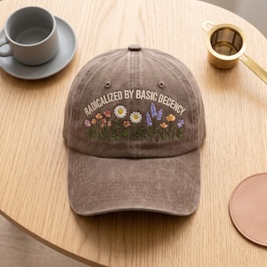 May include: A brown baseball cap featuring embroidered colourful wildflowers and the text "RADICALIZED BY BASIC DECENCY" in white capital letters across the front. The cap has a curved brim and a structured crown with visible stitching details.
