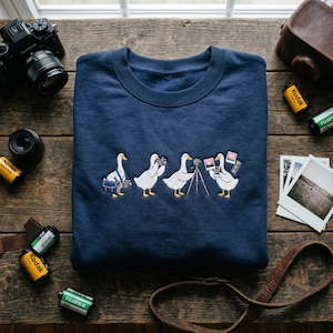 May include: Navy blue sweatshirt featuring an embroidered design of four geese in various photography poses. The geese are holding cameras, with one using a tripod. Film rolls, a camera, and a leather case are also visible.