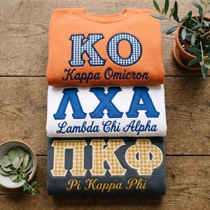 Peut inclure: Trois sweat-shirts à col rond empilés verticalement. Le haut est orange avec des lettres KO en vichy bleu et blanc et le texte Kappa Omicron. Le milieu est blanc avec des lettres bleues ΛXA et le texte Lambda Chi Alpha. Le bas est gris foncé avec des lettres ΠKΦ en vichy jaune et le texte Pi Kappa Phi.