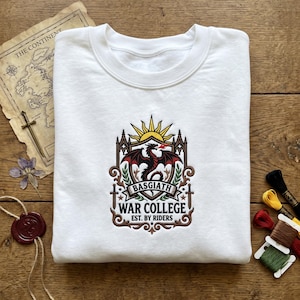 May include: White sweatshirt with a detailed emblem featuring a red dragon, sun, and text that reads "BASGIATH WAR COLLEGE EST. BY RIDERS." The design is in shades of red, brown, and gold. The sweatshirt is folded on a wooden surface.