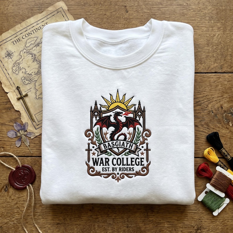 May include: White sweatshirt with a detailed emblem featuring a red dragon, sun, and text that reads "BASGIATH WAR COLLEGE EST. BY RIDERS." The design is in shades of red, brown, and gold. The sweatshirt is folded on a wooden surface.