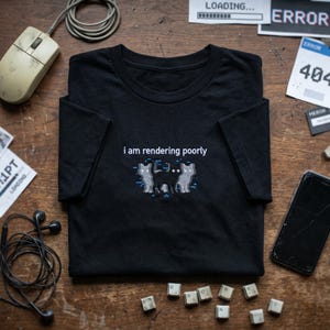 May include: A black short-sleeve t-shirt featuring an embroidered graphic of a central black cat flanked by two grey glitch-effect cats. Above the cats, the white text reads, "i am rendering poorly." The design includes small blue pixelated accents.