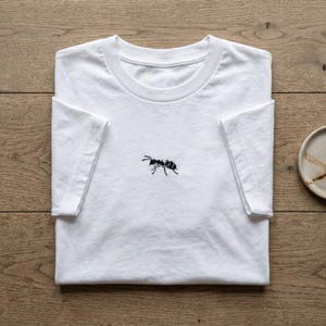May include: White t-shirt featuring a black ant design embroidered on the front. The short-sleeved shirt is folded on a wooden surface. A small, round, ceramic plate is visible in the background.