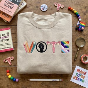 May include: A beige crewneck sweatshirt featuring the embroidered word VOTE. The letter V is formed by three books, the O is a black raised fist symbol, the T is a pink uterus illustration, and the E is a rainbow pride flag pattern.
