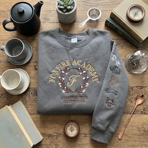 May include: Gray sweatshirt with "FOXFIRE ACADEMY" embroidered in gold, featuring a laurel wreath design and the text "KEEPER OF THE LOST CITIES". The sleeve displays embroidered animal emblems. A teapot, cups, and books surround the sweatshirt.