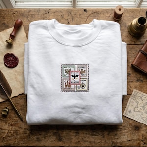 May include: A white crew neck sweatshirt with a colourful stamp design. The stamps feature illustrations of a dragon, a compass, and text including "War College", "Basgiath", and "4th Wing". The sweatshirt is folded on a wooden surface.