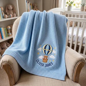 May include: A light blue knitted baby blanket featuring a textured waffle pattern and a ribbed border. The blanket is embroidered with a brown teddy bear in a blue and yellow hot air balloon, white clouds, yellow stars, and the name Oliver James.
