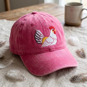 May include: A pink baseball cap with an embroidered rooster design. The rooster has white, yellow, and red details. The cap is made of a soft, washed fabric and has a curved brim.