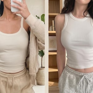May include: Two white ribbed tank tops shown in a side-by-side comparison. The left tank top features wide shoulder straps and a scoop neckline. The right tank top features a high crew neckline and racerback-style armholes. Both are fitted.