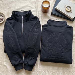 May include: A black quarter-zip sweatshirt featuring tonal embroidery. The front displays a hooded figure on the right sleeve and moon phases, an eye, and text on the left sleeve. The back features a mountain landscape, stars, and the text "VELARIS CITY OF STARLIGHT" and "to the stars who listen and the dreams that are answered."