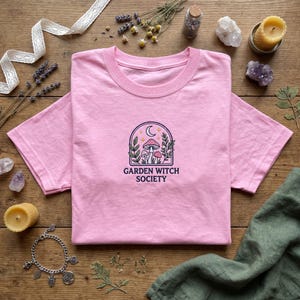 Könnte beinhalten: Hellrosa T-Shirt mit Rundhalsausschnitt. Das Shirt zeigt ein gesticktes Design aus Pilzen, Blättern, einem Halbmond und Sternen in einem bogenförmigen Rahmen. Der Text "GARDEN WITCH SOCIETY" befindet sich darunter.