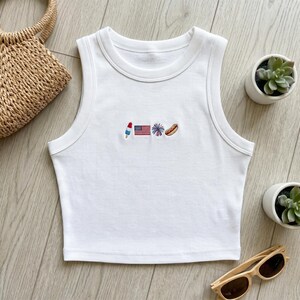 May include: A white ribbed tank top featuring a small embroidered design on the center chest. The embroidery consists of a red, white, and blue popsicle, an American flag, a blue and red firework burst, and a brown hot dog on a bun.