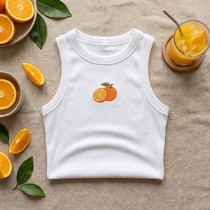 May include: A white ribbed tank top featuring a small embroidered graphic of a whole orange and a sliced orange half with a green leaf in the center. The garment has a crew neckline and sleeveless design, showcasing a clean and simple aesthetic.