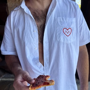 May include: A white short-sleeved button-down shirt featuring a chest pocket with an embroidered red heart outline containing the letters D&R. The shirt is made of a lightweight fabric and is worn unbuttoned, revealing a plain white collar and front.