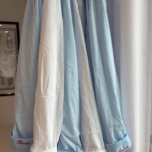 May include: A collection of long-sleeved button-down shirts in alternating light blue and white colors. Each shirt features a folded cuff embroidered with a unique name in red script, including the names Tiana, Lauren, Hannah, and Kate.
