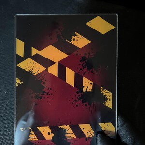 May include: A rectangular acrylic display case featuring a dark red background with black ink splatters and diagonal yellow and black hazard warning stripes printed across the surface. The case has a clear, smooth finish and a protective frame.