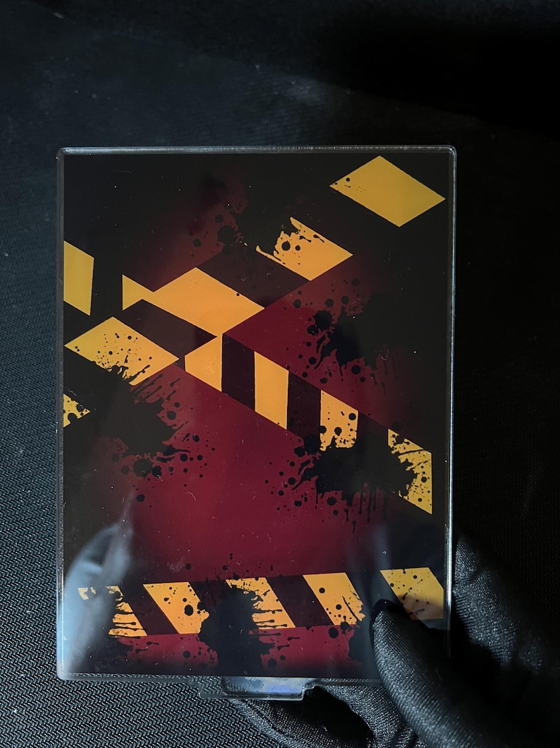 May include: A rectangular acrylic display case featuring a dark red background with black ink splatters and diagonal yellow and black hazard warning stripes printed across the surface. The case has a clear, smooth finish and a protective frame.