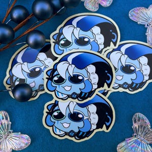 May include: A collection of die-cut stickers featuring a stylized blue-skinned character with large dark eyes, white hair, and a dark blue hood. The character has a small white frilled collar and a dark blue emblem at the neck, depicted in a cartoon style.