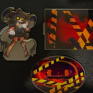 May include: An acrylic standee featuring a cartoon character with yellow X-shaped eyes, a rectangular acrylic plate with orange and black hazard stripes, and an oval acrylic base with matching hazard stripes and red paint splatter patterns.