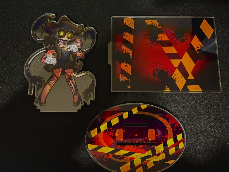 May include: An acrylic standee featuring a cartoon character with yellow X-shaped eyes, a rectangular acrylic plate with orange and black hazard stripes, and an oval acrylic base with matching hazard stripes and red paint splatter patterns.