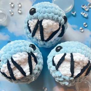 Puede incluir: Tres juguetes redondos de ganchillo azul hechos a mano con detalles blancos y negros. Cada juguete tiene ojos negros ovalados y una amplia sonrisa con rayas negras. Los juguetes están colocados sobre un fondo azul claro con gemas transparentes dispersas.