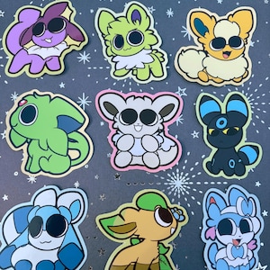 May include: A collection of nine die-cut stickers featuring stylized cartoon creatures with large black eyes. The characters include various color palettes such as purple, lime green, yellow, light green, white, black, blue, and tan with distinct features.
