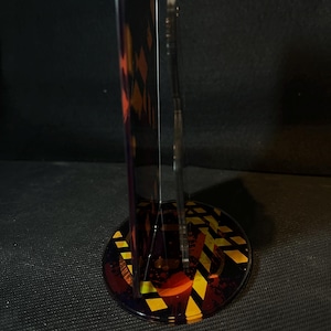 May include: A vertical acrylic display stand featuring a circular base with a yellow and black hazard stripe pattern. The stand includes a transparent upright panel and a secondary vertical piece, both decorated with dark, abstract graphic elements.