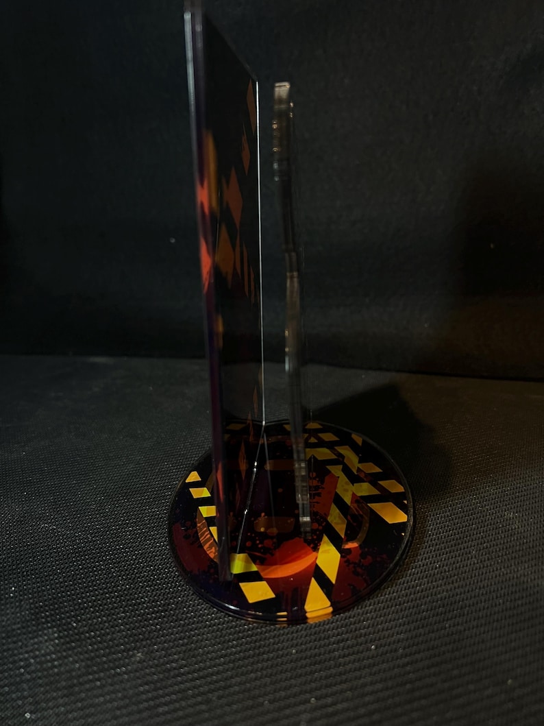 May include: A vertical acrylic display stand featuring a circular base with a yellow and black hazard stripe pattern. The stand includes a transparent upright panel and a secondary vertical piece, both decorated with dark, abstract graphic elements.