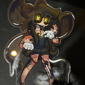 May include: A clear acrylic standee featuring a cartoon character with long brown hair, glowing yellow X-shaped eyes, and a wide grin. The character wears a dark dress with a skull pendant, white gloves, and has dark, ink-like stains on their skin.