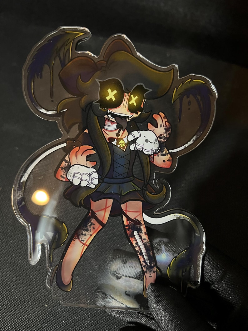 May include: A clear acrylic standee featuring a cartoon character with long brown hair, glowing yellow X-shaped eyes, and a wide grin. The character wears a dark dress with a skull pendant, white gloves, and has dark, ink-like stains on their skin.