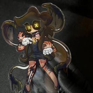 May include: A clear acrylic standee featuring a stylized character with long brown hair, yellow X-shaped eyes, and a dark dress. The character has white gloves, dark stockings with red grid patterns, and dark flowing tendrils with purple accents.