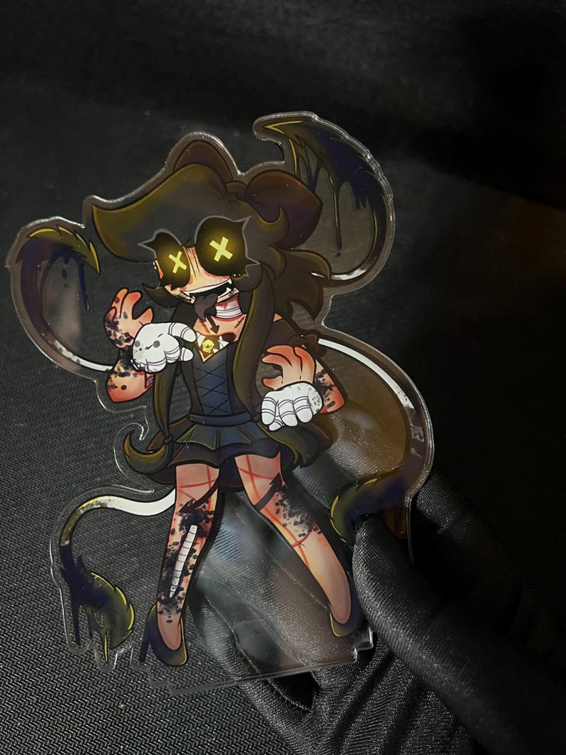 May include: A clear acrylic standee featuring a stylized character with long brown hair, yellow X-shaped eyes, and a dark dress. The character has white gloves, dark stockings with red grid patterns, and dark flowing tendrils with purple accents.