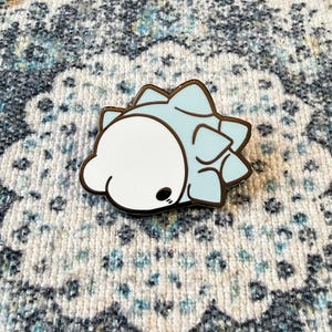 May include: A metal enamel pin shaped like a stylized creature with a smooth white rounded head and a light blue spiky body. The design features dark brown outlines and a small black eye detail on the white portion of the character.