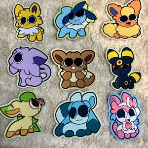 May include: A collection of nine die-cut stickers featuring stylized cartoon creatures with large black eyes. The characters include various color palettes such as yellow, blue, orange, purple, brown, black, green, and pink, each with distinct shapes.