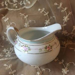 May include: A white porcelain creamer with a light blue rim and floral design. The creamer has a delicate floral pattern with pink and blue flowers.