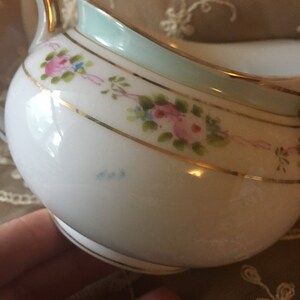 May include: A white porcelain creamer with a light blue rim and gold trim. The creamer is decorated with hand-painted pink roses and blue flowers.