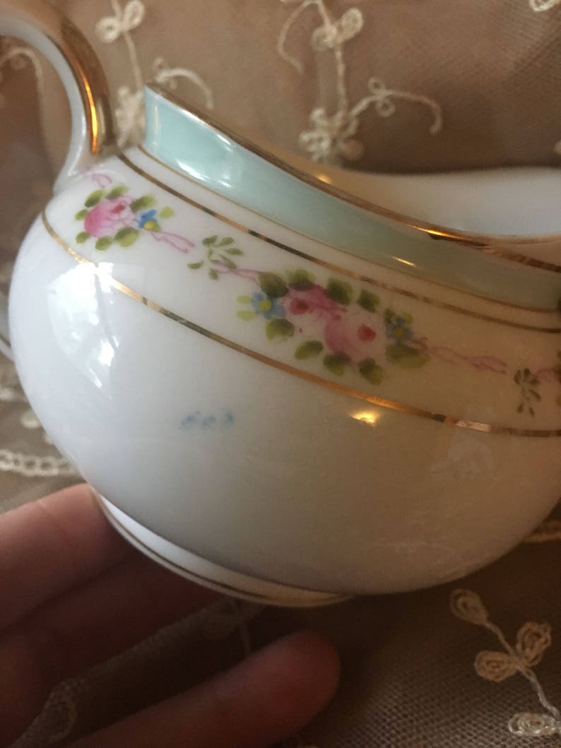 May include: A white porcelain creamer with a light blue rim and gold trim. The creamer is decorated with hand-painted pink roses and blue flowers.