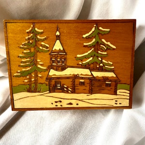 May include: A wooden box with a carved scene of a snow-covered cabin with a church steeple in the background. The cabin is surrounded by two pine trees and the scene is painted in shades of brown, green, and white.
