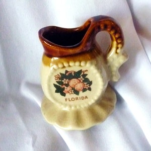 May include: A small brown and white ceramic creamer with a ruffled base and a handle. It features a design of oranges and leaves with the word "FLORIDA" printed on the front.