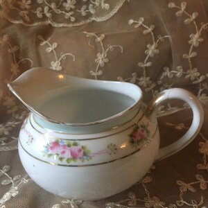 May include: A white porcelain creamer with a delicate floral design in pink, green, and gold. The creamer has a light blue band around the rim and a gold band around the base.