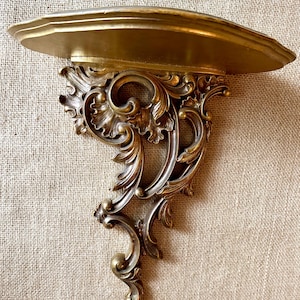 May include: A wall-mounted shelf featuring a semi-circular top surface and an ornate, decorative bracket underneath. The entire piece is finished in a metallic gold tone with intricate, carved-style scrollwork and leaf-like detailing.