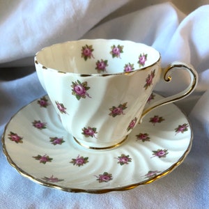 May include: A white teacup and saucer set with a delicate pink rose pattern. The cup and saucer have a gold trim around the edges.