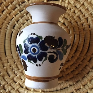 May include: A small white ceramic vase with a blue and brown floral design. The vase has a rounded body and a narrow neck.