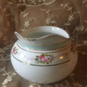 May include: A white porcelain creamer with a delicate floral design featuring pink roses and green leaves. The creamer has a gold trim and a light blue band around the rim.