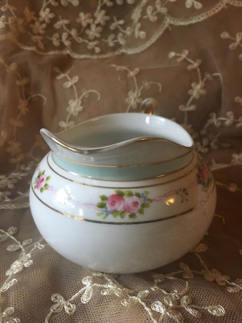 May include: A white porcelain creamer with a delicate floral design featuring pink roses and green leaves. The creamer has a gold trim and a light blue band around the rim.