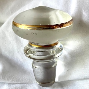 Może przedstawiać: Dekoracyjny korek do butelki z matową białą górą i złotą opaską. Korek ma przezroczystą szklaną podstawę i zwężane dno. Projekt jest prosty i elegancki, odpowiedni do różnych butelek.