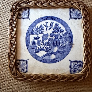 May include: Square decorative tray with a woven wicker frame. The center features a blue and white porcelain design with a landscape scene. The corners have blue floral accents. The tray is set on a neutral-colored surface.