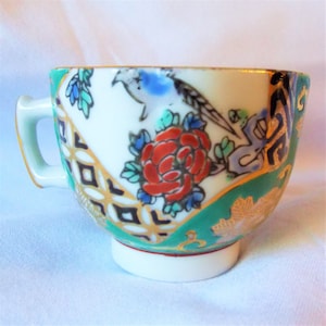 May include: A small, white porcelain teacup with a green, gold, and blue floral design. The cup has a handle and a rounded base.