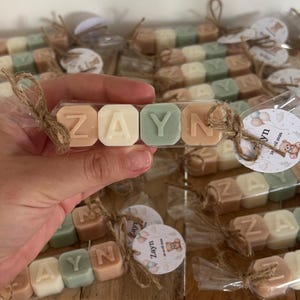 Peut inclure: Quatre blocs carrés de couleur brun clair, crème et vert sauge, chacun présentant une lettre en relief pour épeler ZAYN. Les blocs sont emballés dans un plastique transparent noué avec de la ficelle et une étiquette ronde avec un ours.